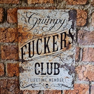 May include: A white metal sign with a distressed, rusty look. The sign reads "Grumpy Fuckers Club" with the words "Lifetime Member" below. The sign has a crown and a skull and crossbones design.