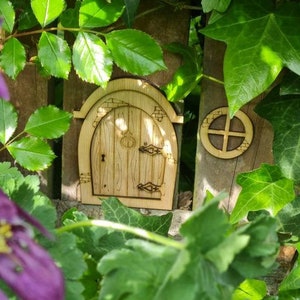 May include: A wooden fairy door with a rounded archway and a small window, nestled in a bed of green leaves and purple flowers.