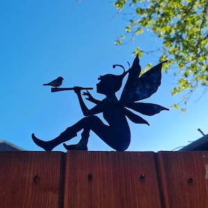May include: A black metal silhouette of a fairy with wings sitting on a wooden fence, playing a flute and looking at a small bird.