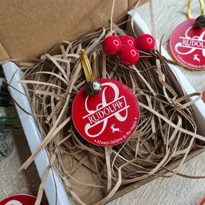 May include: A red and white personalized Christmas ornament with a bell and the name "Rudolph" and the text "Always believe in the magic." The ornament is in a box with red and white tissue paper.