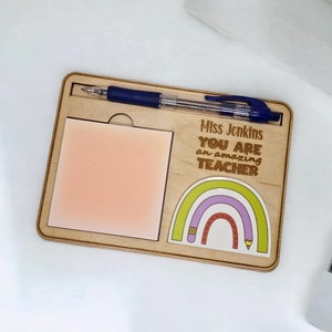 Op de afbeelding: Houten bureau-organizer met een notitieblok, een pennenhouder en een gepersonaliseerd bericht dat luidt "Miss Jenkins YOU ARE an amazing TEACHER". Het bericht wordt vergezeld door een regenboog met een potlood.