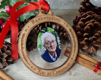 Może przedstawiać: Drewniana ozdoba świąteczna z napisem "Remembering You at Christmas" otaczającym zdjęcie starszej osoby. Ozdoba ma czerwoną wstążkę do zawieszenia i jest otoczona szyszkami i laskami cynamonu.