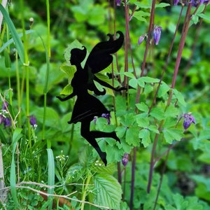 Könnte beinhalten: Schwarze Metall-Fee-Silhouette mit ausgestreckten Armen und detaillierten Flügeln vor dem Hintergrund von üppigem grünem Laub und lila Blumen. Die Fee ist ein dekoratives Gartenelement.