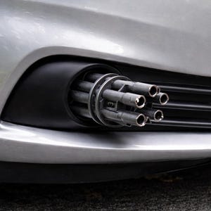Op de afbeelding: De voorkant van een zilveren auto, met een zwarte behuizing in de bumper die een multi-loop geweer bevat. De lopen van het geweer zijn zichtbaar en de grille van de auto is zwart.
