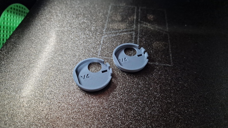 Pode incluir: Dois pequenos componentes circulares azul claro impressos em 3D com um orif&iacute;cio central e duas ranhuras. Cada componente tem o n&uacute;mero "14" impresso. Os componentes est&atilde;o sobre uma superf&iacute;cie escura texturizada.