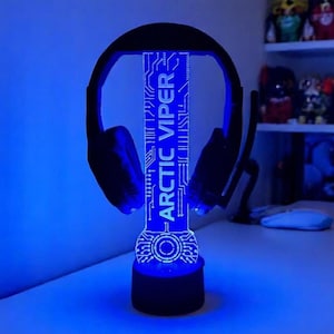 Op de afbeelding: Een zwarte headset rust op een blauw verlichte standaard. De standaard toont de tekst "ARCTIC VIPER" in het wit, met een printplaatontwerp. De basis en de headset zijn zwart, wat een coole, technisch geïnspireerde uitstraling creëert.