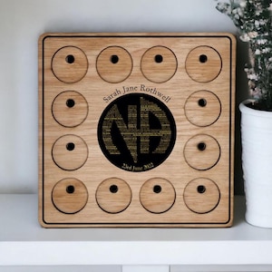 May include: A wooden plaque with 12 circles, each with a small hole in the center. The plaque has a black border and a personalized inscription that reads "Sarah Jane Rothwell NA 23rd June 2022".