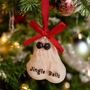 May include: A novelty Christmas tree ornament shaped like a pair of testicles, with the words "Jingle Balls" printed on it. The ornament is cream-colored, has two black bells at the top, and is suspended by a red ribbon.