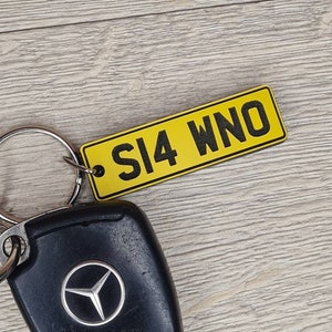 May include: A yellow keychain with black text that reads "SI4 WNO" attached to a black car key with a Mercedes-Benz logo.