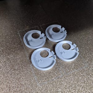Pode incluir: Quatro pe&ccedil;as cinzentas, circulares, impressas em 3D, com um furo central e um entalhe. Cada pe&ccedil;a tem o texto "V4" impresso. As pe&ccedil;as est&atilde;o dispostas numa superf&iacute;cie castanha texturizada.