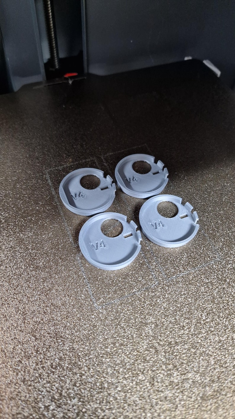 Pode incluir: Quatro pe&ccedil;as cinzentas, circulares, impressas em 3D, com um furo central e um entalhe. Cada pe&ccedil;a tem o texto "V4" impresso. As pe&ccedil;as est&atilde;o dispostas numa superf&iacute;cie castanha texturizada.