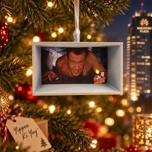 May include: A rectangular Christmas ornament featuring a scene of a person in a vent holding a lit lighter. The frame is light blue with a textured border. A hanging tag attached to the ornament displays the handwritten text Yippee Ki Yay.
