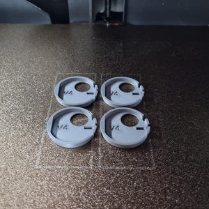 Pode incluir: Quatro objetos circulares cinza claro, impressos em 3D, com um orif&iacute;cio central e uma borda entalhada, est&atilde;o dispostos sobre uma superf&iacute;cie texturizada marrom escuro. Os objetos parecem ser componentes ou pe&ccedil;as de um item maior.