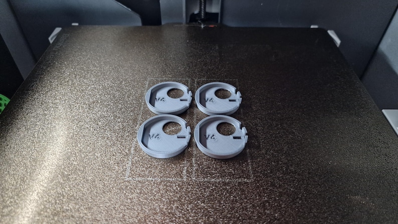 Pode incluir: Quatro objetos circulares cinza claro, impressos em 3D, com um orif&iacute;cio central e uma borda entalhada, est&atilde;o dispostos sobre uma superf&iacute;cie texturizada marrom escuro. Os objetos parecem ser componentes ou pe&ccedil;as de um item maior.