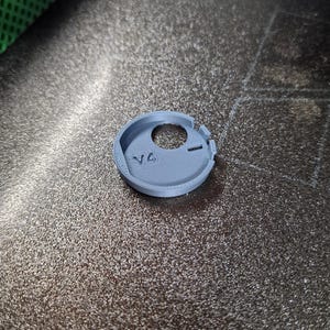 Pode incluir: Um objeto circular cinza claro, impresso em 3D, com um orif&iacute;cio central e um pequeno entalhe. O objeto tem as letras "V4" impressas em sua superf&iacute;cie. O item est&aacute; sobre uma superf&iacute;cie escura texturizada.