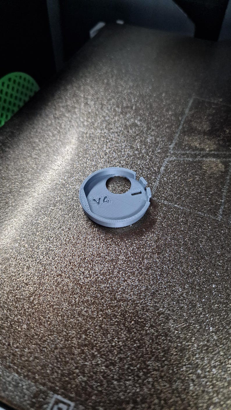 Pode incluir: Um objeto circular cinza claro, impresso em 3D, com um orif&iacute;cio central e um pequeno entalhe. O objeto tem as letras "V4" impressas em sua superf&iacute;cie. O item est&aacute; sobre uma superf&iacute;cie escura texturizada.