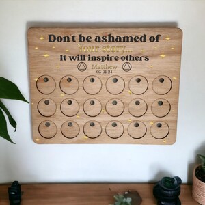 Könnte beinhalten: Holztafel mit dem Text "Schäm dich nicht für deine Geschichte... Sie wird andere inspirieren" und dem Datum "05-01-21". Die Tafel hat 24 runde Ausschnitte.