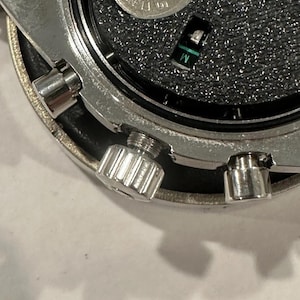 Pode incluir: Close-up dos componentes internos de um rel&oacute;gio. A imagem mostra uma caixa de metal prateada com um interior preto texturizado. Uma pequena bateria e um componente verde e preto s&atilde;o vis&iacute;veis. O rel&oacute;gio tem uma coroa aparafusada e outras pequenas pe&ccedil;as de metal.