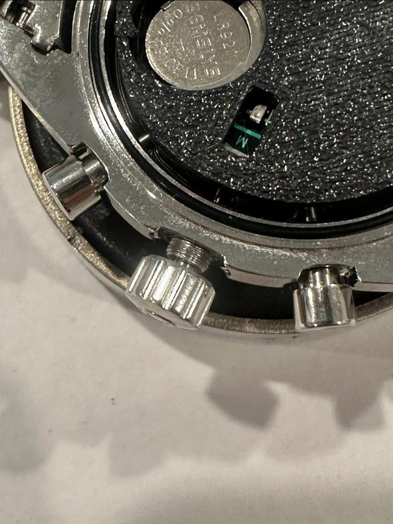 Pode incluir: Close-up dos componentes internos de um rel&oacute;gio. A imagem mostra uma caixa de metal prateada com um interior preto texturizado. Uma pequena bateria e um componente verde e preto s&atilde;o vis&iacute;veis. O rel&oacute;gio tem uma coroa aparafusada e outras pequenas pe&ccedil;as de metal.