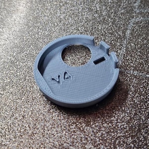 Pode incluir: Um objeto circular cinza claro, impresso em 3D, com um furo central e dois pequenos recortes retangulares. As letras "VA" s&atilde;o vis&iacute;veis. Parece ser um pequeno componente.