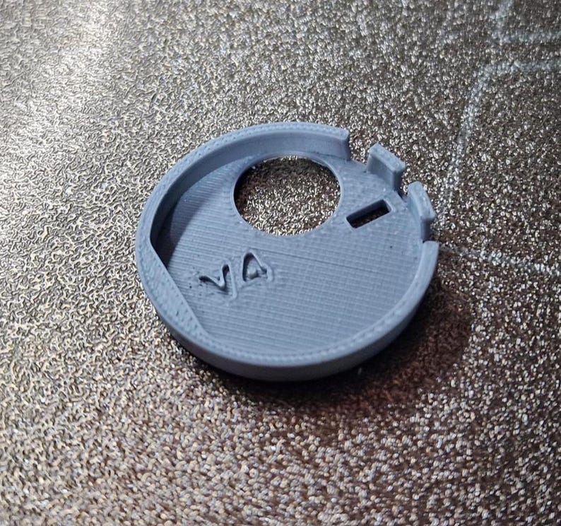 Pode incluir: Um objeto circular cinza claro, impresso em 3D, com um furo central e dois pequenos recortes retangulares. As letras "VA" s&atilde;o vis&iacute;veis. Parece ser um pequeno componente.