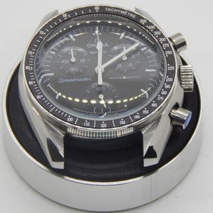 Pode incluir: Um rel&oacute;gio de pulso preto e prateado com uma luneta taquim&eacute;trica. O mostrador do rel&oacute;gio exibe a marca "Omega" e o texto "Speedmaster Moonwatch". O rel&oacute;gio est&aacute; sobre um suporte preto e prateado.