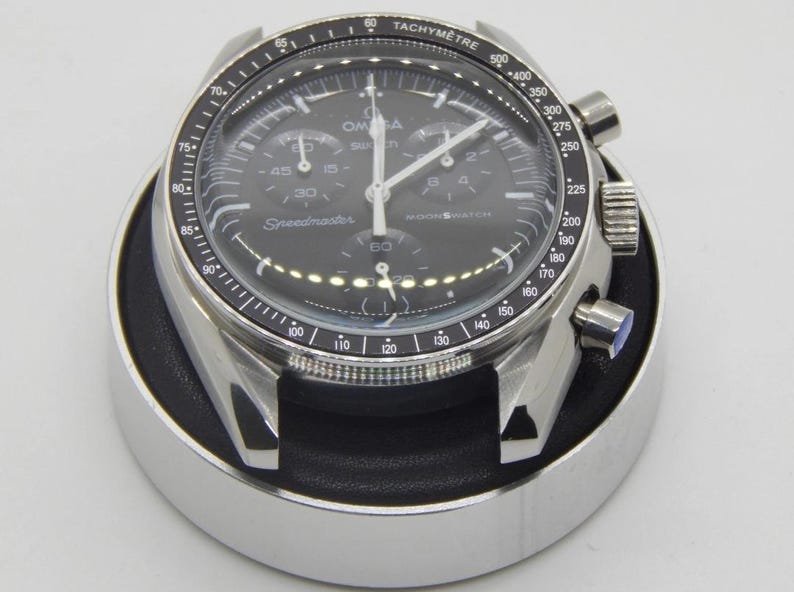 Pode incluir: Um rel&oacute;gio de pulso preto e prateado com uma luneta taquim&eacute;trica. O mostrador do rel&oacute;gio exibe a marca "Omega" e o texto "Speedmaster Moonwatch". O rel&oacute;gio est&aacute; sobre um suporte preto e prateado.