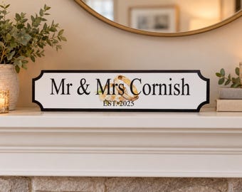 May include: A rectangular white sign with a black border featuring the text Mr & Mrs Cornish in black serif font. Centered between the words is an illustration of two gold wedding bands intertwined with white flowers and the text EST. 2025 below.