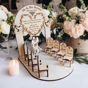 May include: A wooden wedding ceremony set with a heart-shaped arch, two doves, and a couple standing beneath it. The set includes six miniature chairs with personalized names, including "Father of the Bride", "Aunt Maureen", "Janet", and "Joanna".