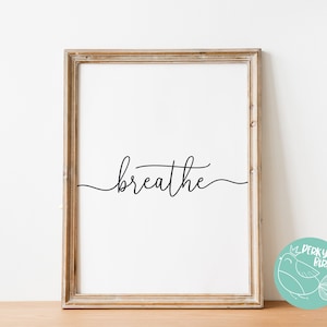 May include: A framed print with the word "breathe" in elegant cursive script. The artwork is set against a white background, framed in a light brown wooden frame. A teal circular logo with a bird is in the lower right corner.