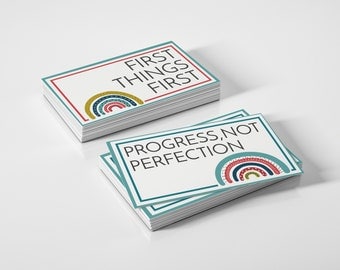 Puede incluir: Dos pilas de tarjetas rectangulares con fondos blancos. La tarjeta superior de una pila dice "FIRST THINGS FIRST" en texto negro, y la otra "PROGRESS, NOT PERFECTION". Cada tarjeta presenta un diseño de arcoíris.