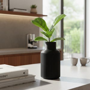 Puede incluir: Un jarrón negro acanalado con una planta verde sobre una encimera blanca. El jarrón tiene un diseño de rayas verticales. El fondo muestra una cocina con luz natural y dos tazas.