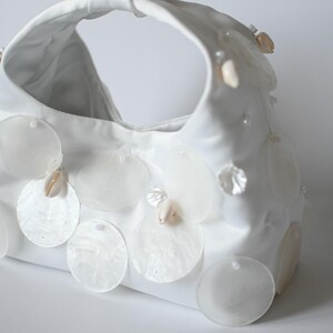 May include: A white satin handbag decorated with large, round, iridescent shell embellishments and small seashells. The bag has a curved shape with a single handle. Ideal for a beach wedding or summer occasion.