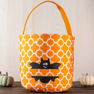 May include: Orange and white patterned Halloween treat bag with a black bat design. The bag has an orange handle and is surrounded by small pumpkins. A festive container for trick-or-treating.