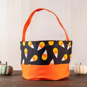 May include: A Halloween-themed candy corn tote bag with an orange handle and base. The main body of the bag is black with a repeating candy corn pattern. The bag is sitting on a wooden surface with decorative pumpkins.