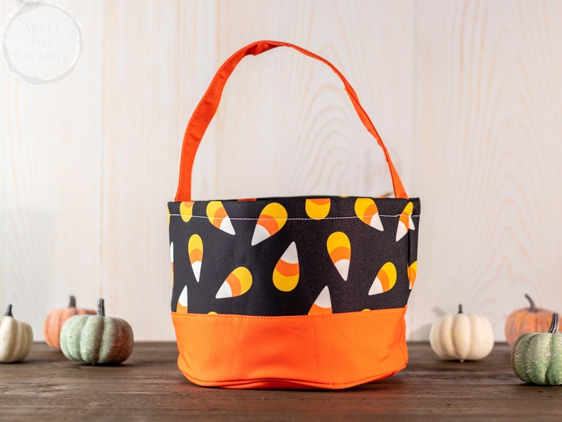 May include: A Halloween-themed candy corn tote bag with an orange handle and base. The main body of the bag is black with a repeating candy corn pattern. The bag is sitting on a wooden surface with decorative pumpkins.