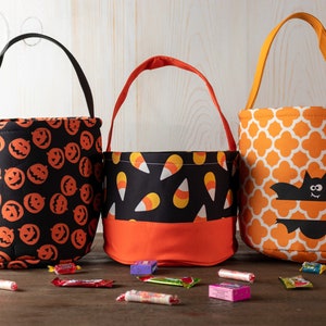 May include: Three Halloween-themed fabric treat bags. One black bag with orange jack-o'-lantern faces, a second with candy corn, and a third with an orange and white quatrefoil pattern and a bat applique. Black, red, and orange handles.