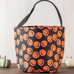 May include: Black Halloween bucket bag with orange jack-o'-lantern faces. The bag has a black handle and is made of fabric. The bucket is sitting on a wooden surface with decorative pumpkins in the background.