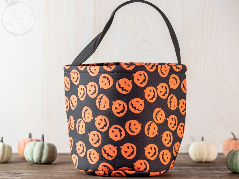 May include: Black Halloween bucket bag with orange jack-o'-lantern faces. The bag has a black handle and is made of fabric. The bucket is sitting on a wooden surface with decorative pumpkins in the background.