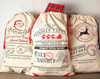 May include: Three cream-colored Christmas gift bags with red drawstrings and festive designs. One bag features Santa Claus, another says "Reindeer Express," and the third says "Reindeer Mail." A plaid gift box sits atop the bags, adding a touch of holiday cheer.
