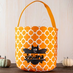 May include: Orange and white patterned Halloween treat bag with a black bat graphic and the name "Juliana". The bag has an orange handle and is surrounded by small pumpkins.