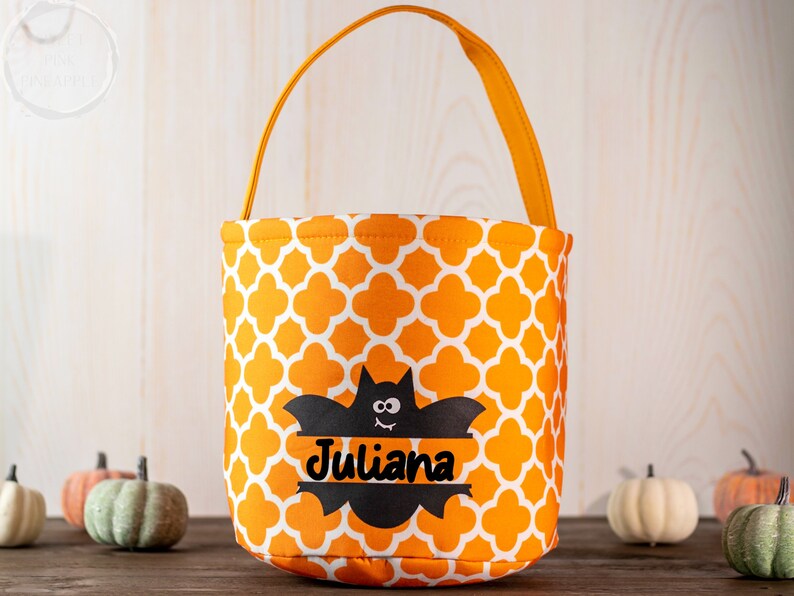 May include: Orange and white patterned Halloween treat bag with a black bat graphic and the name "Juliana". The bag has an orange handle and is surrounded by small pumpkins.