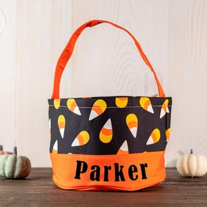 May include: A Halloween candy bag with an orange handle and base, and a black upper section patterned with candy corn. The name "Parker" is printed in black on the orange base. Small decorative pumpkins are in the background.