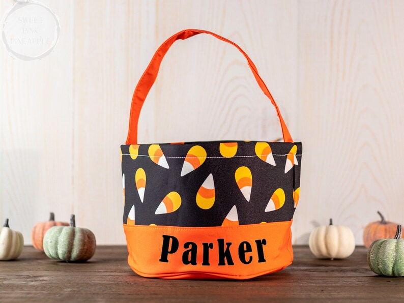 May include: A Halloween candy bag with an orange handle and base, and a black upper section patterned with candy corn. The name "Parker" is printed in black on the orange base. Small decorative pumpkins are in the background.