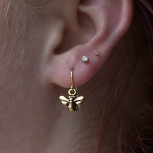 May include: A close-up of a person's ear with three small earrings. The bottom earring is a gold bee charm dangling from a gold hoop. The other two earrings are small, clear gemstones.