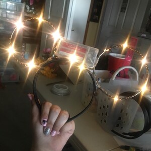 Puede incluir: Un par de orejas de diadema negras con un marco de alambre dorado y luces LED blancas. Las luces est&aacute;n encendidas y brillan.