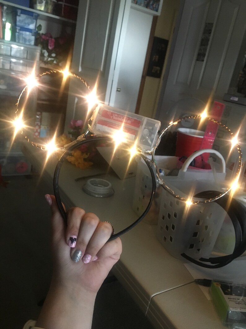 Puede incluir: Un par de orejas de diadema negras con un marco de alambre dorado y luces LED blancas. Las luces est&aacute;n encendidas y brillan.