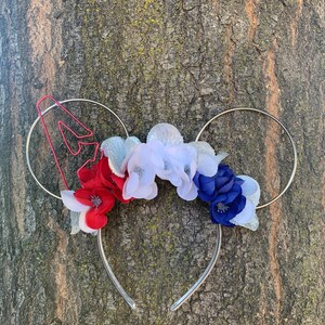 Pode incluir: Flores de tecido vermelho, branco e azul presas a uma faixa de cabeça prateada com um 4 de metal vermelho de um lado.