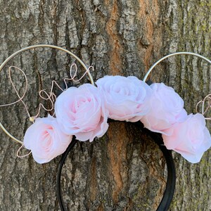 May include: A pair of black mouse ears with a silver wire headband. The headband is decorated with pink fabric roses and the word "Love" written in silver wire.