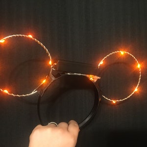 Puede incluir: Una diadema negra con dos c&iacute;rculos de luz naranja unidos a ella. Los c&iacute;rculos est&aacute;n hechos de alambre y tienen peque&ntilde;as luces naranjas incrustadas.
